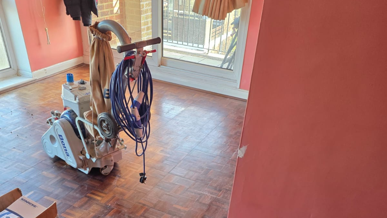 Wood Floor Sanding in Honor Oak on a hardwood mosaic parquet floor, showing a Bona belt sander preparing the worn surface in a bright living room.