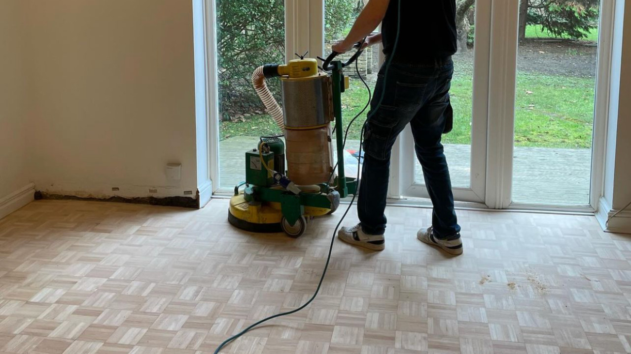 Wood Floor Sanding in Honor Oak on a light hardwood mosaic finger parquet floor, with a technician using a large rotary sander near patio doors overlooking the garden.