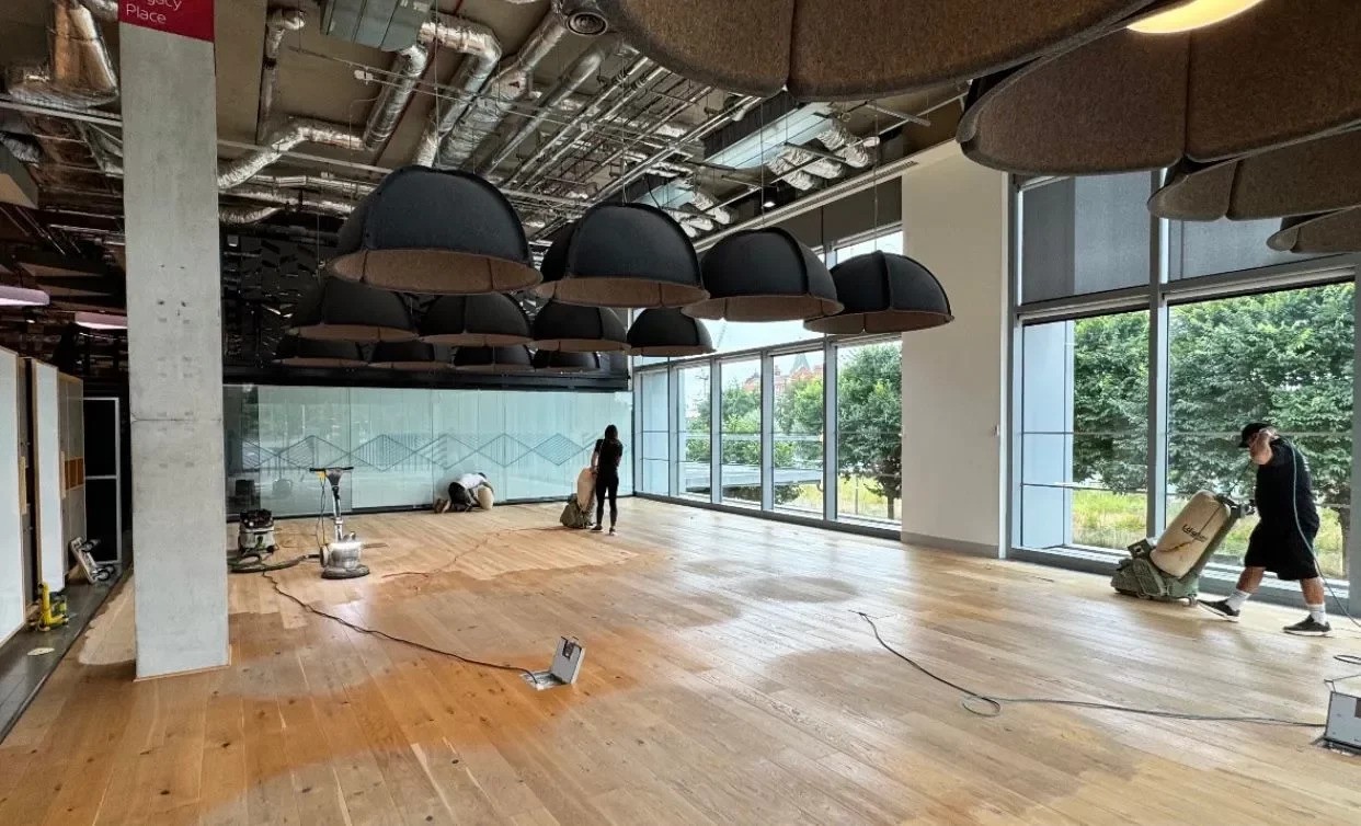Wood Floor Sanding in Honor Oak on an oak wide-plank hardwood office floor, with technicians using sanding machines in a large, modern workspace with full-height windows.