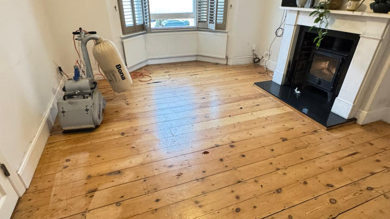 Wood Floor Sanding in Honor Oak on pine softwood floorboards, showing a living room being sanded with a Bona belt sander near a bay window and fireplace.